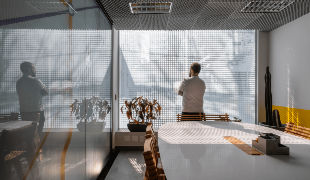 Imagem mostra homem de pele branca de costas em um escritório de coworking, olhando pela janela