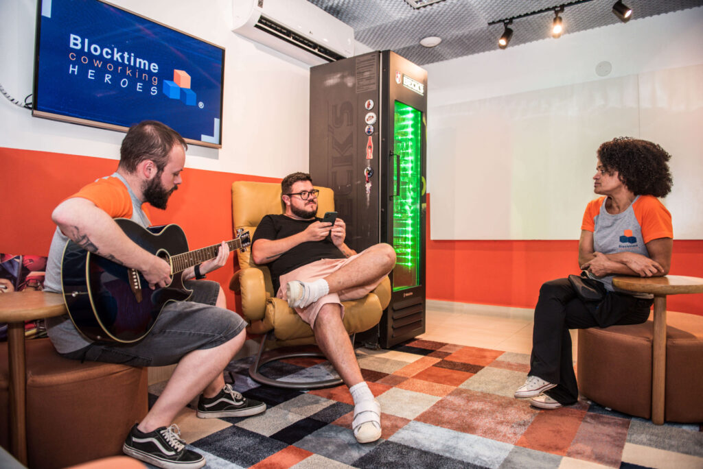 Imagem mostra pessoas descontraídas, sentadas em um ambiente da Blocktime Coworking, conversando e tocando violão