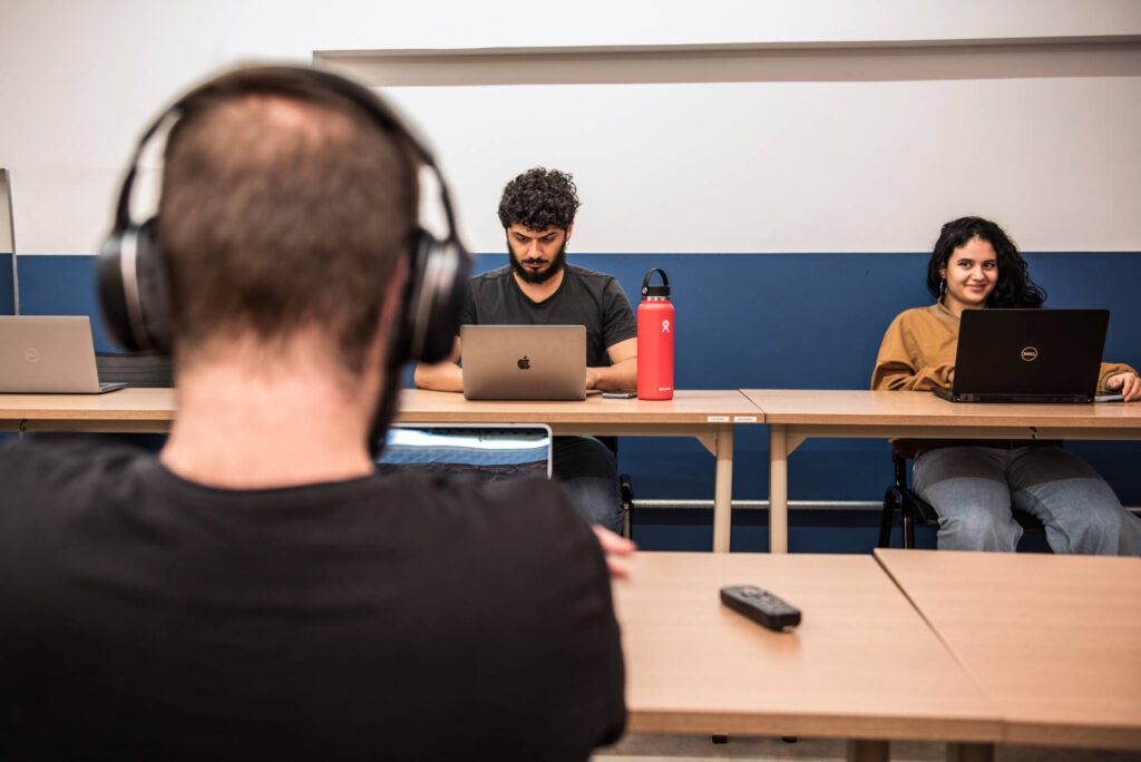 Imagem mostra três pessoas trabalhando com computadores em um coworking. Um está de costas para a imagem e os outros dois estão de frente, olhando para o computador.