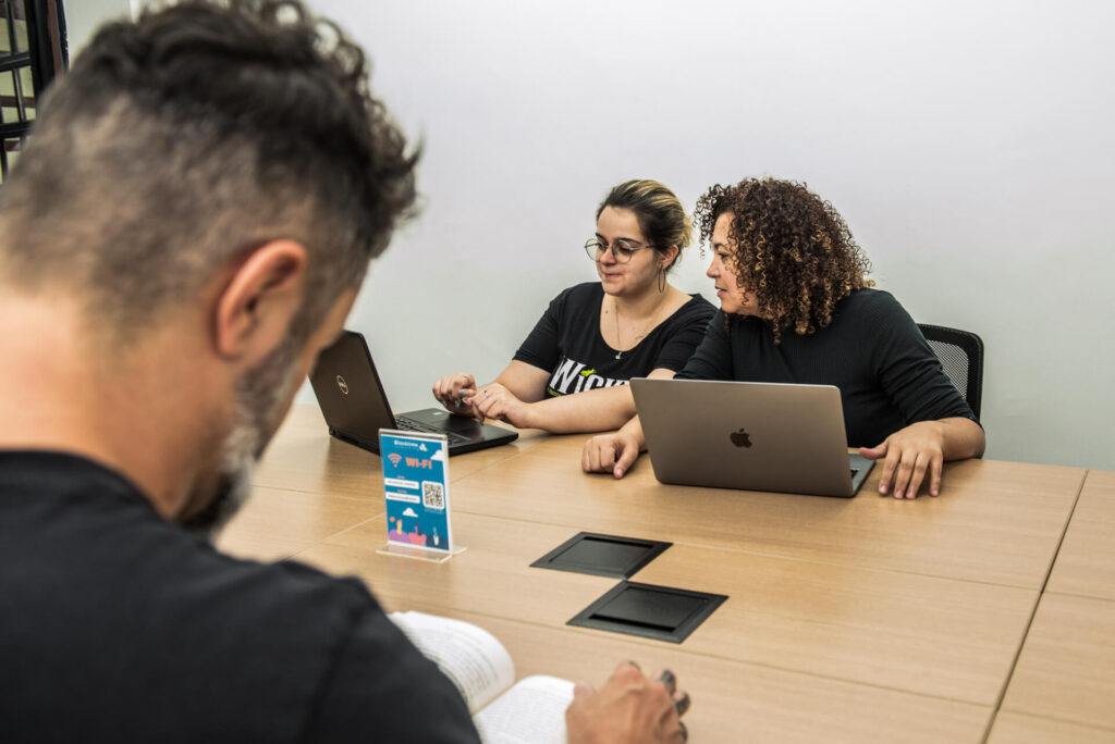 Imagem mostra três pessoas trabalhando com computadores em um coworking. Um está de costas para a imagem e os outros dois estão olhando para o computador e conversando.
