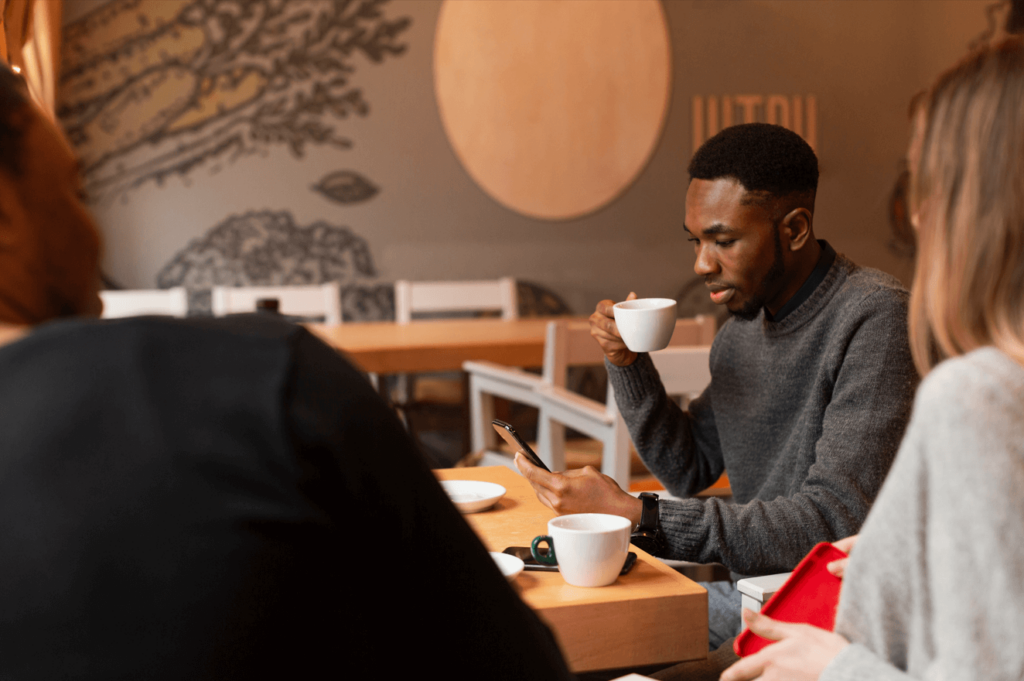 Imagem mostra um homem jovem, preto, tomando um café e olhando para o seu celular