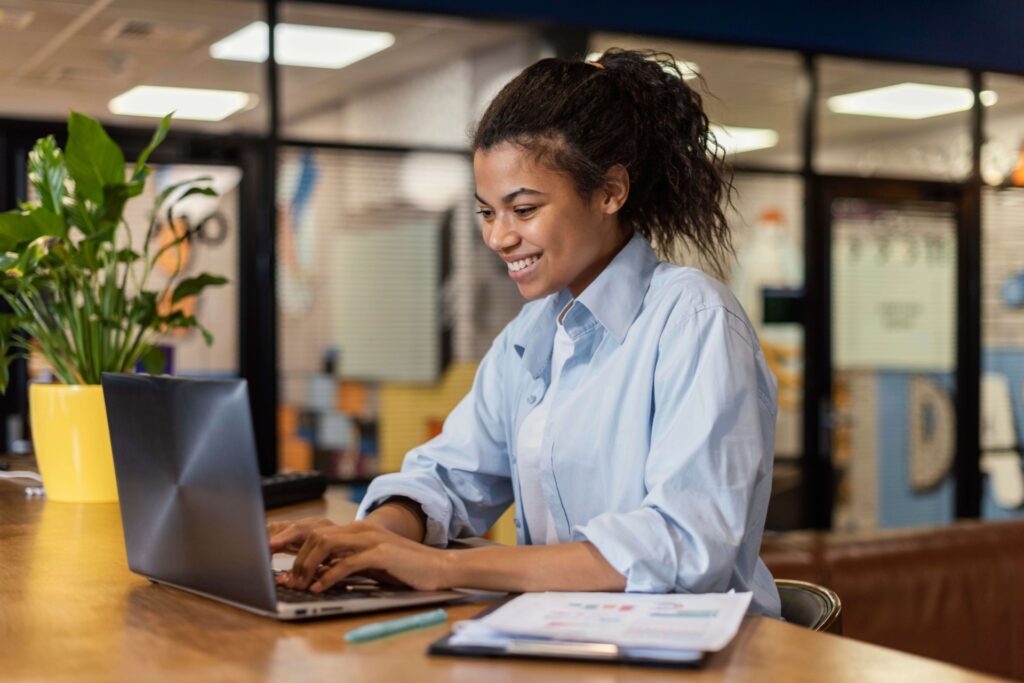 A imagem mostra uma mulher jovem preta, sentada numa mesa de escritório, trabalhando no computador e sorrindo