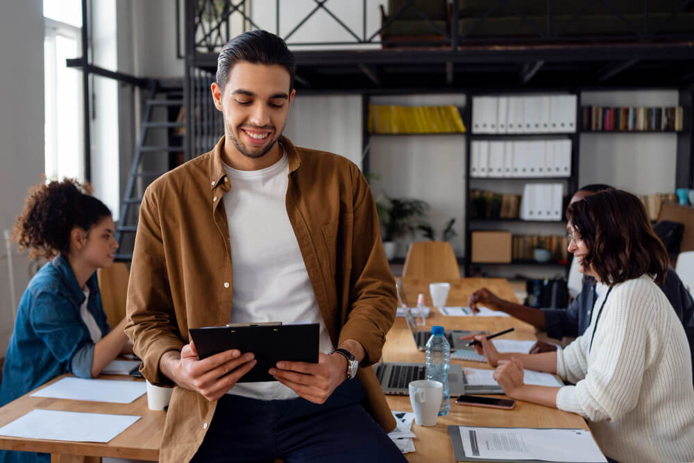 Imagem mostra homem sentado na mesa, segurando tablet e ao fundo está seu time de trabalho sentado nas cadeiras