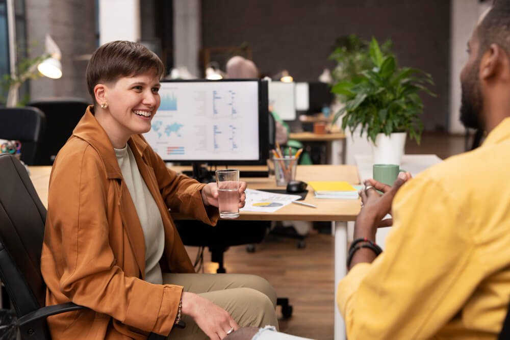 Imagem mostra pessoas em ambiente de trabalho, estão sentada conversando, uma olhando para a outra.