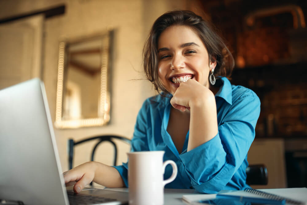 Imagem mostra uma mulher de pele clara, jovem, sentada a mesa, com mão no teclado do notebook e sorrindo e olhando para a camera.