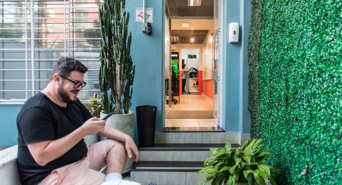 Imagem mostra homem jovem sentado na entrada da Blocktime Coworking, na frente dele há uma parede com folhas verticais.