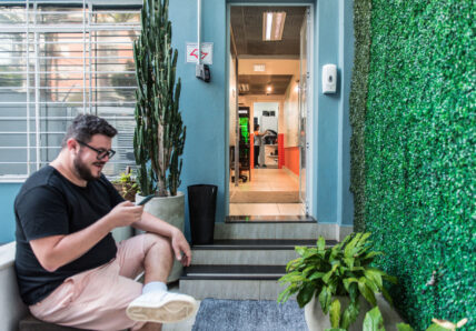 Imagem mostra homem jovem sentado na entrada da Blocktime Coworking, na frente dele há uma parede com folhas verticais.
