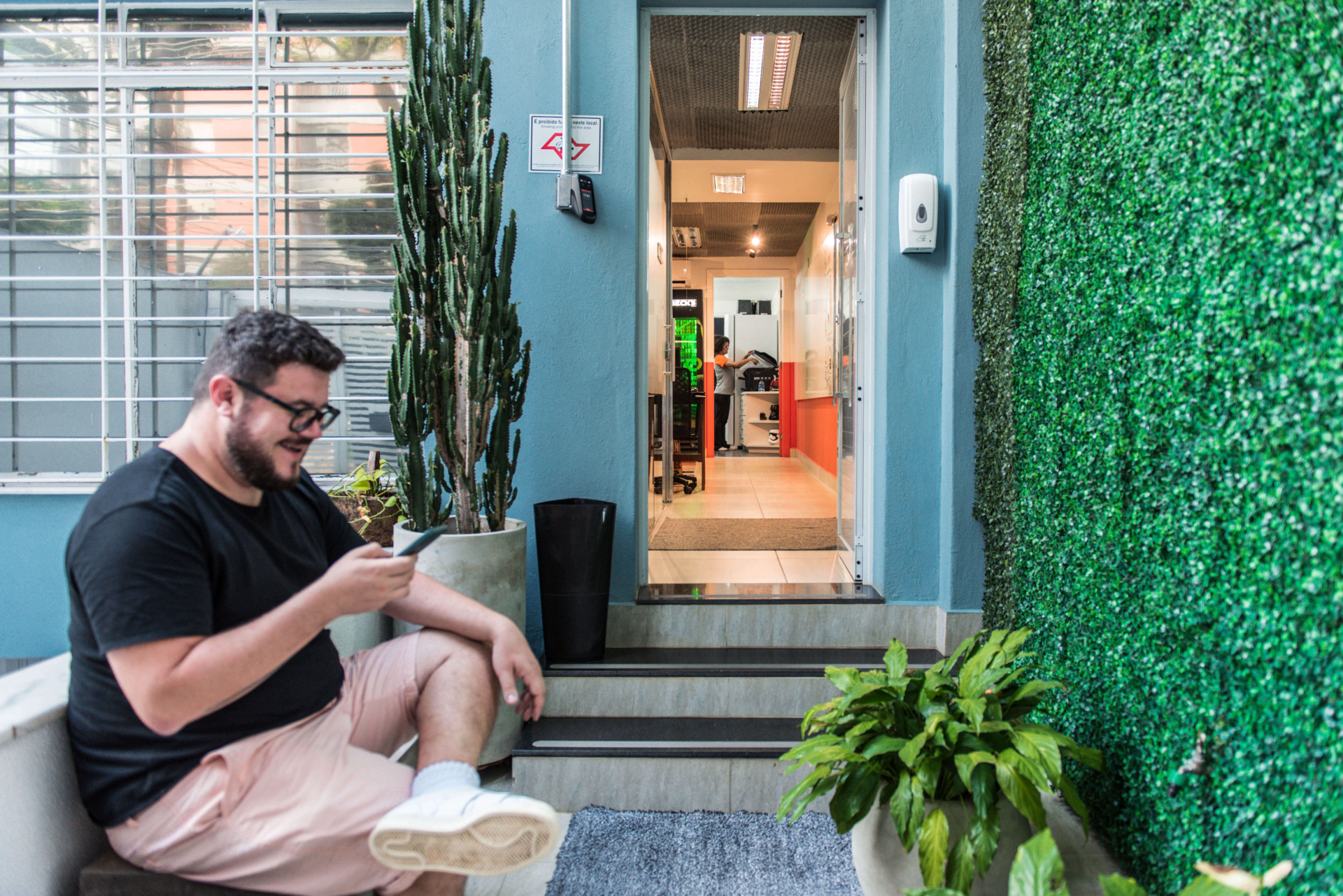 Imagem mostra homem jovem sentado na entrada da Blocktime Coworking, na frente dele há uma parede com folhas verticais.