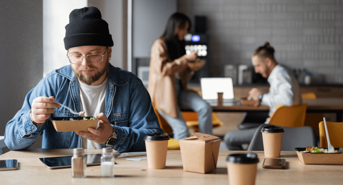 Imagem mostra um homem branco, jovem, sentado na mesa de um café com pessoas ao fundo conversando, enquanto ele come