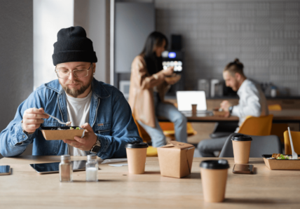 Imagem mostra um homem branco, jovem, sentado na mesa de um café com pessoas ao fundo conversando, enquanto ele come