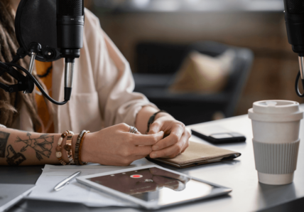 Imagem mostra mulher sentada numa mesa de escritório. Em cima da mesa tem um tablet e um copo de café. E na imagem, não aparece o rosto dela. Ela está gravando um podcast.