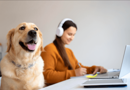 Imagem mostra uma moça jovem trabalhando, usando fones de ouvido e seu cachorro ao lado dela na mesa