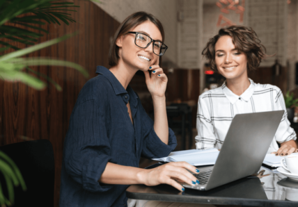 Imagem mostra duas mulheres jovens conversando e rindo enquanto olham para o computador