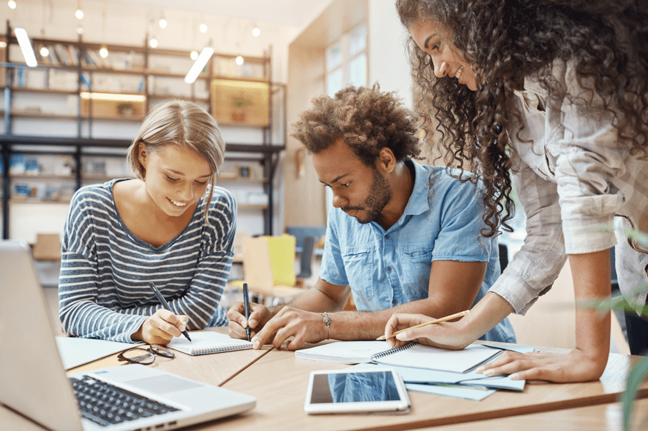Imagem mostra grupo de pessoas diversas, com 2 mulheres e 1 homem, em uma mesa de escritório trabalhando
