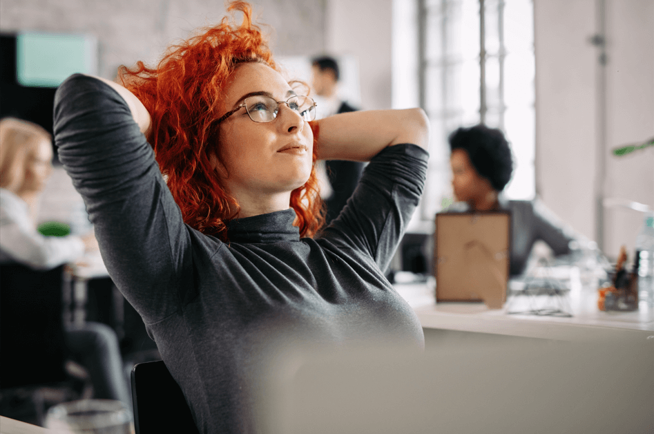 Imagem mostra mulher jovem, de pele clara e cabelos vermelhos, sentada no escritório com os braços na cabeça, passando a sensação de estar relaxada.