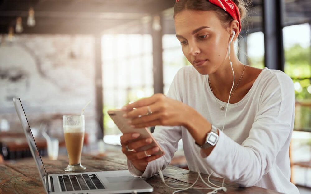 Imagem mostra mulher jovem, de pele clara, sentada em um café, mexendo no celular e na sua frente está seu notebook