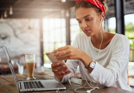 Imagem mostra mulher jovem, de pele clara, sentada em um café, mexendo no celular e na sua frente está seu notebook