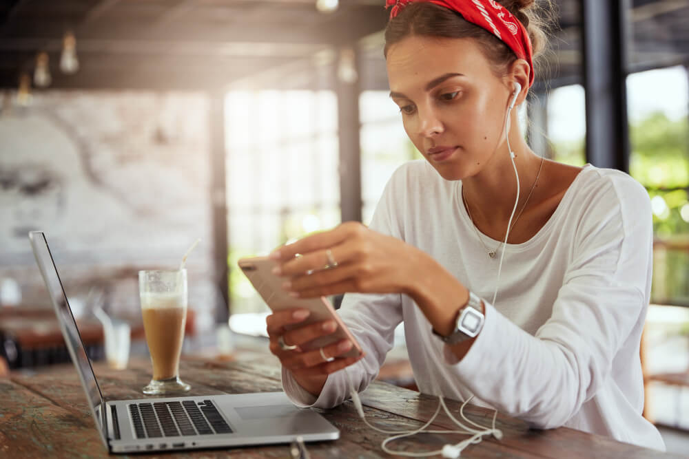 Imagem mostra mulher jovem, de pele clara, sentada em um café, mexendo no celular e na sua frente está seu notebook