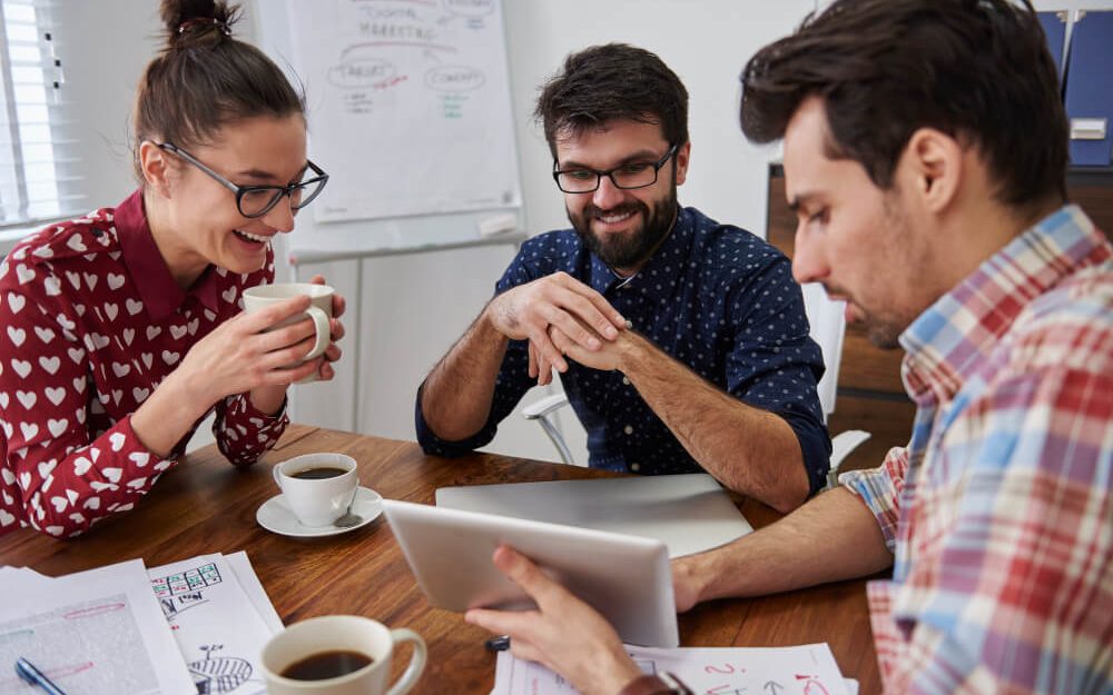 Imagem mostra um grupo de três pessoas sentadas em uma mesa em frente a um computador e cada uma delas está com uma xícara de café