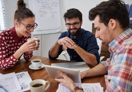 Imagem mostra um grupo de três pessoas sentadas em uma mesa em frente a um computador e cada uma delas está com uma xícara de café