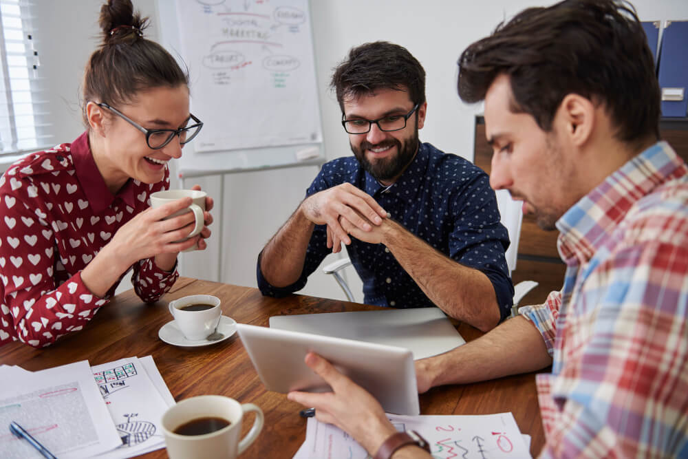 Imagem mostra um grupo de três pessoas sentadas em uma mesa em frente a um computador e cada uma delas está com uma xícara de café