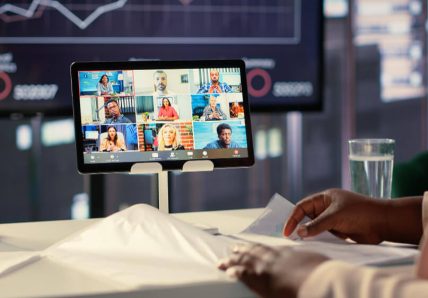 Imagem mostra uma pessoa conectada em reunião online. A Imagem mostra ela sentada de frente pra tela do computador, na tela há várias online em reunião. Ela está fazendo anotações.
