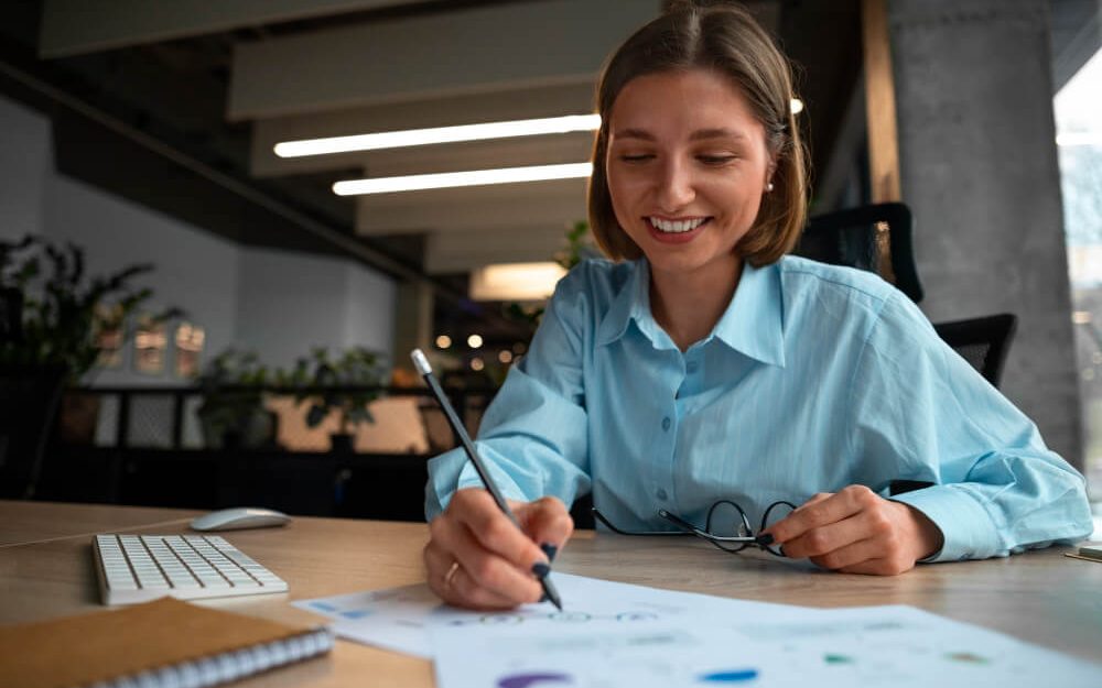 Imagem mostra mulher de pele clara escrevendo e fazendo anotações em papés com gráficos financeiros