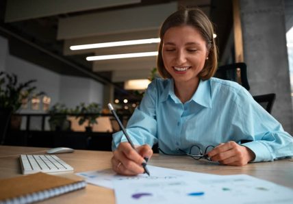 Imagem mostra mulher de pele clara escrevendo e fazendo anotações em papés com gráficos financeiros