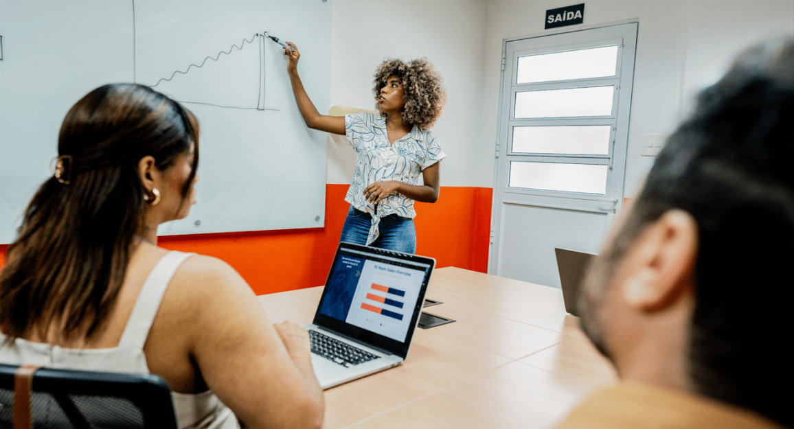 Imagem mostra mulher escrevendo em lousa em uma sala de reunião da Blocktime Coworking, enquanto isso, duas pessoas olham para sua direção, acompanhando o que ela escreve.