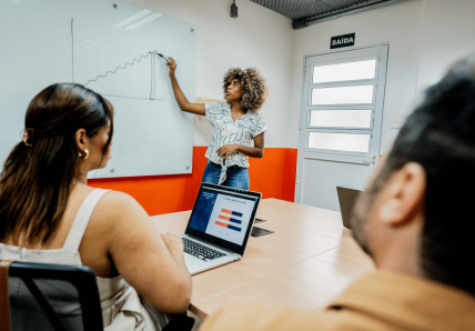 Imagem mostra mulher escrevendo em lousa em uma sala de reunião da Blocktime Coworking, enquanto isso, duas pessoas olham para sua direção, acompanhando o que ela escreve.