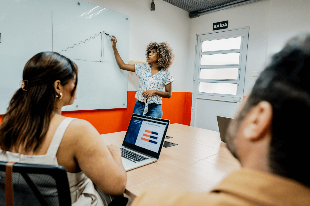 Imagem mostra mulher escrevendo em lousa em uma sala de reunião da Blocktime Coworking, enquanto isso, duas pessoas olham para sua direção, acompanhando o que ela escreve.
